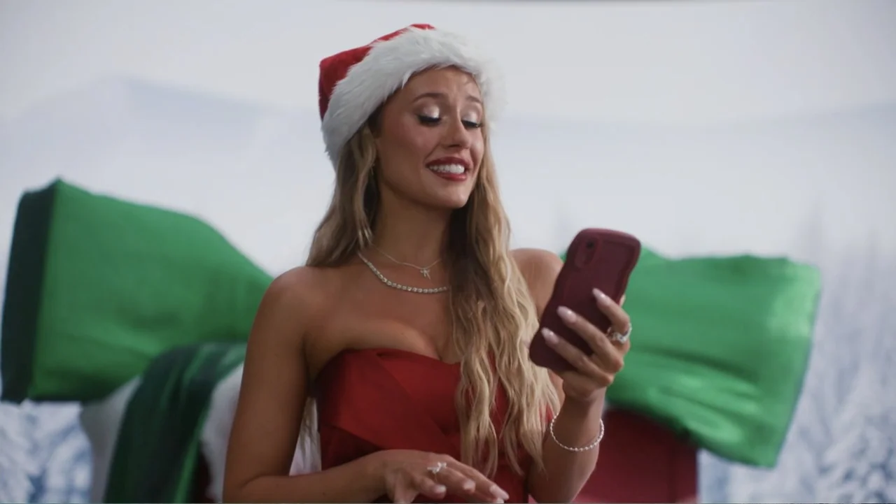 Christmas in the Spotlight (2024) movie still: medium — A studio environment features a large, oversized gift bow in the background. A young woman…