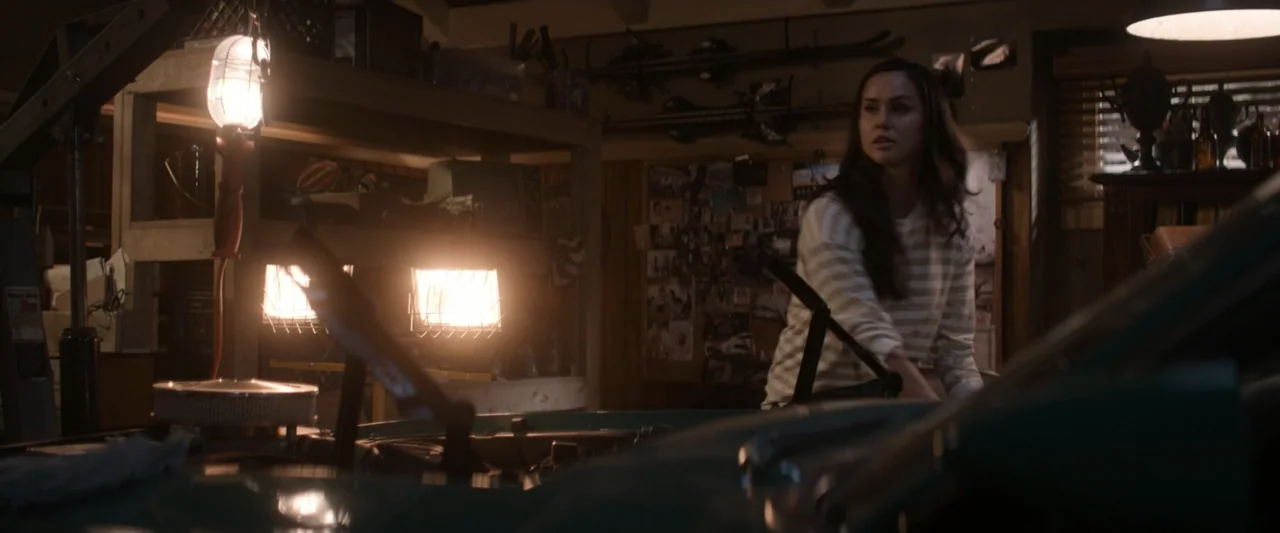On a Wing and a Prayer (2023) movie still: medium — A cluttered garage containing a vintage car with its hood raised and tools stored. A young woman…