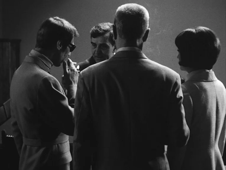 The Rite (1969) movie still: medium — Four individuals gather in a dimly lit interior space with minimalist walls. One man lights a cigarette while…