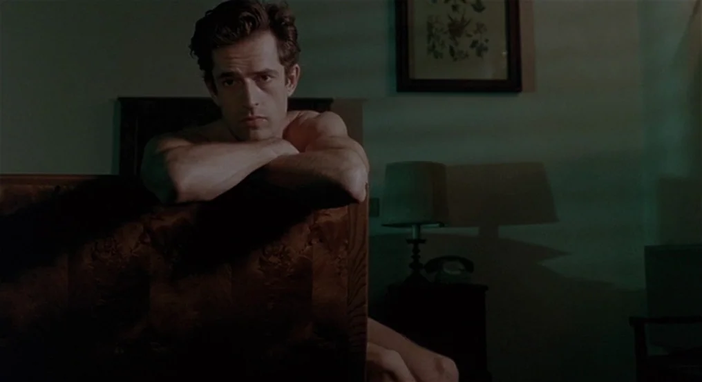The Comfort of Strangers (1990) movie still: medium — A dimly lit room contains a wooden headboard and a bedside lamp. A shirtless man sits behind…