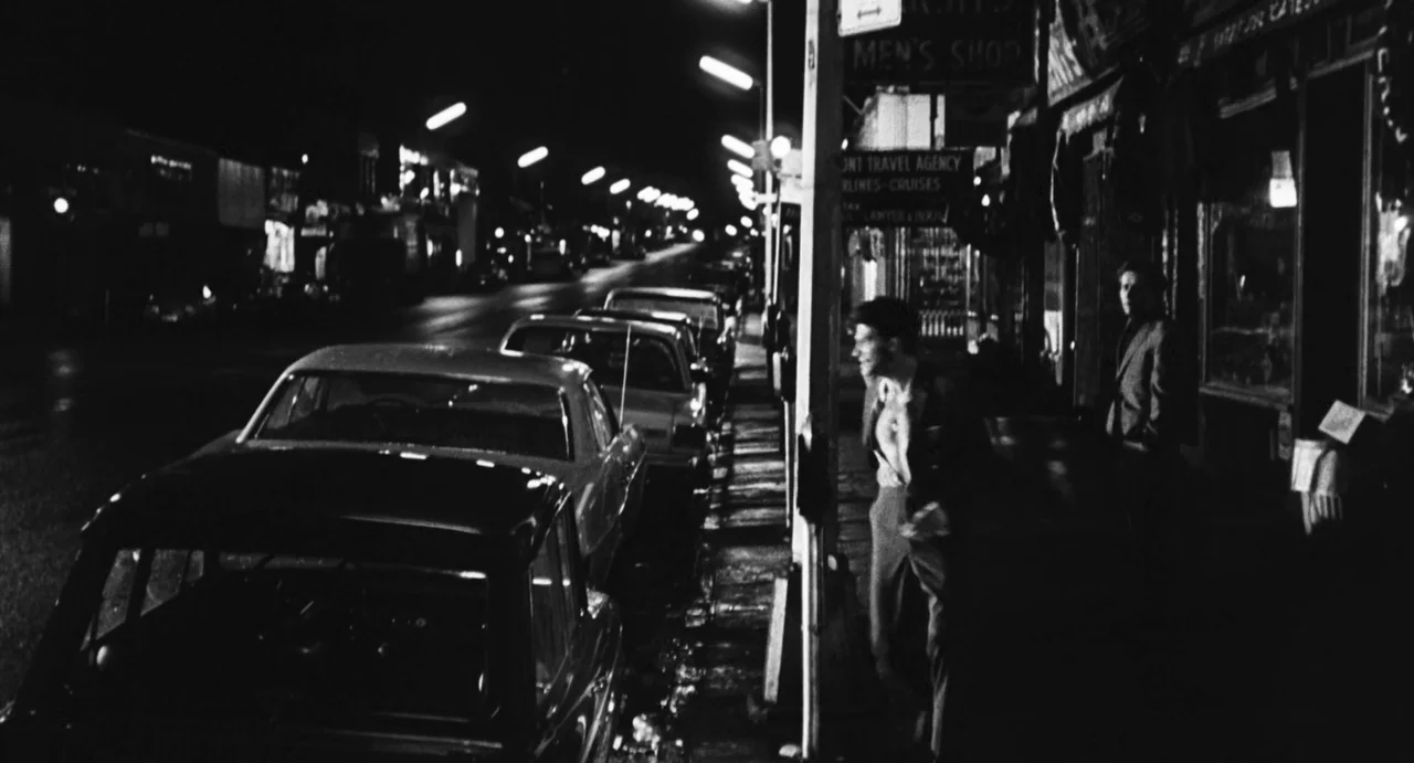 The Incident (1967) movie still: wide — A night street scene features a row of parked cars and storefronts. Two men stand on the sidewalk in the…