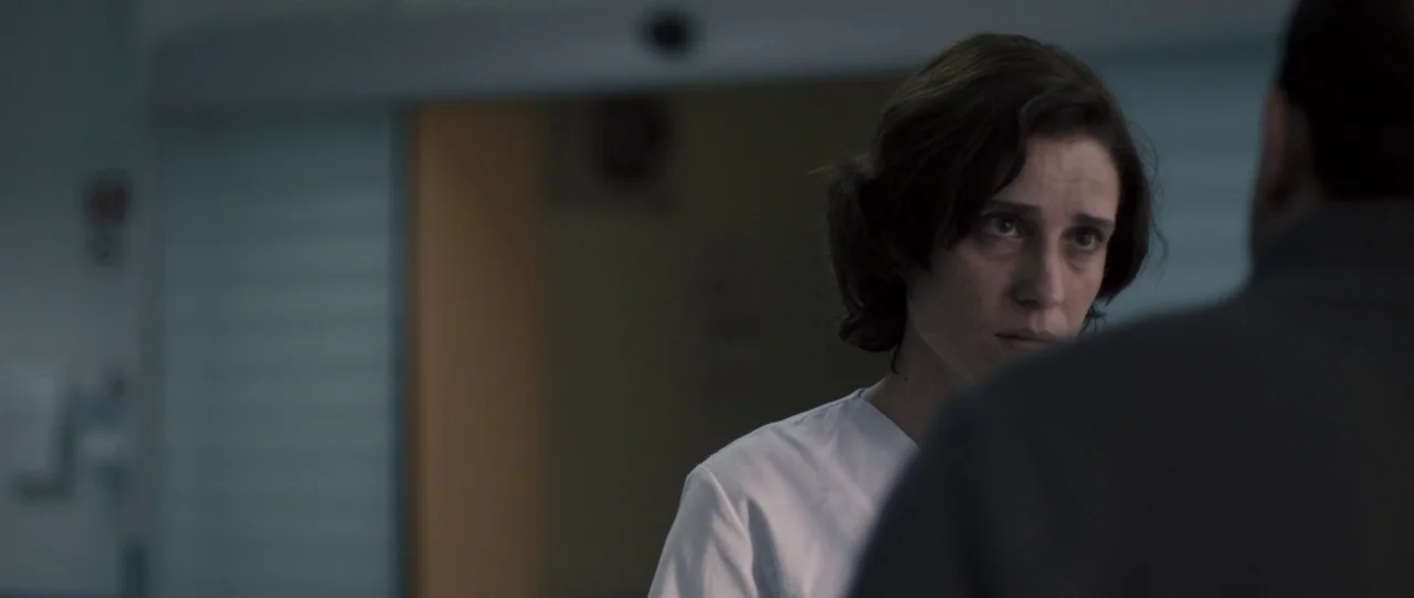 Alps (2011) movie still: medium — A dimly lit hospital corridor with a blurred medical door in the background. A woman in a white scrub top stands…