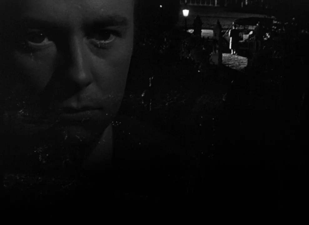 The Magnificent Ambersons (1942) movie still: closeup — A man's face appears as a reflection in a dark, nocturnal window frame. In the distance, a…