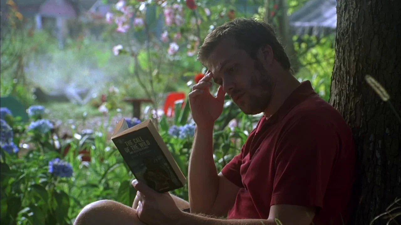 Love Song For Bobby Long (2004) movie still: medium — A man sits beneath a tree in a lush green backyard with blue flowers. He rests his head on his…