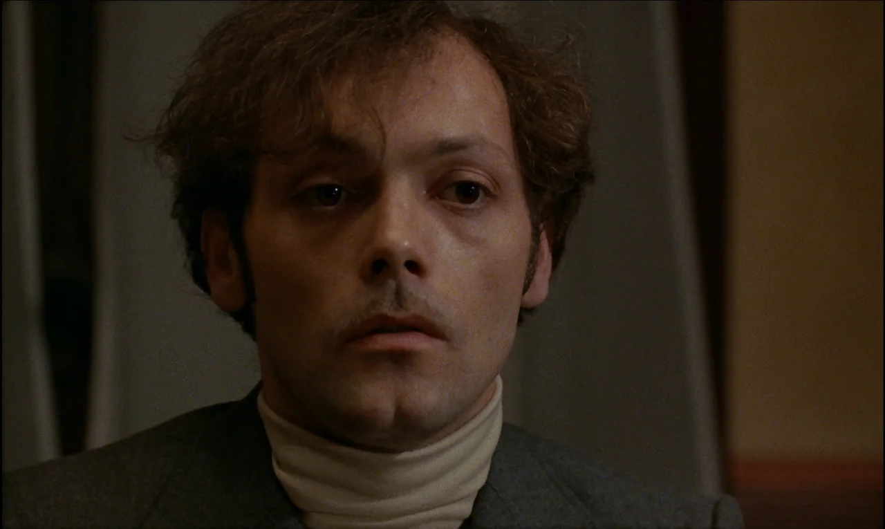 Serie Noire (1979) movie still: closeup — A dimly lit room with draped curtains visible in the background. A man looks directly into the camera with…