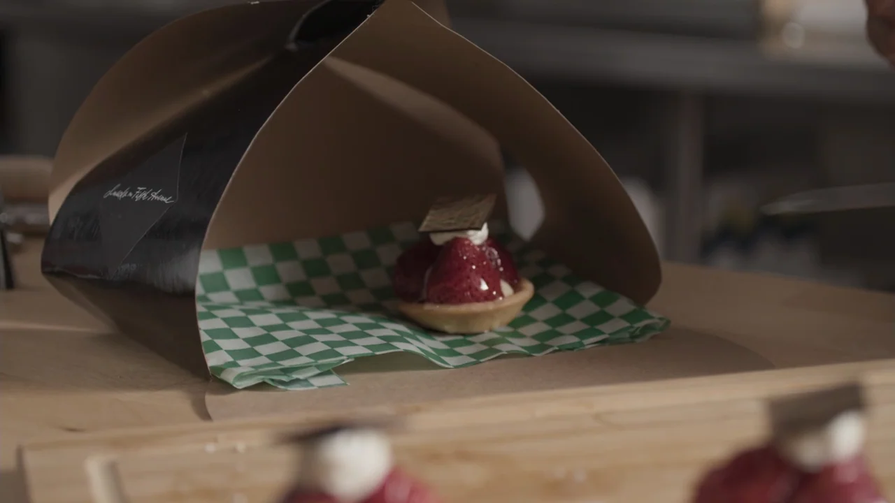 A Gingerbread Christmas (2022) movie still: closeup — A strawberry tart topped with chocolate sits inside a brown cardboard takeout box. The box…