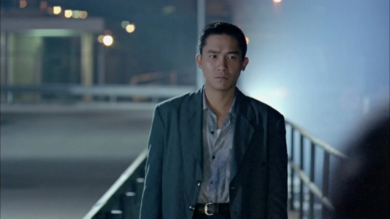 Hard Boiled (1992) movie still: medium — A night view on an outdoor pedestrian walkway with metal guardrails under distant lamps. A man in a dark…