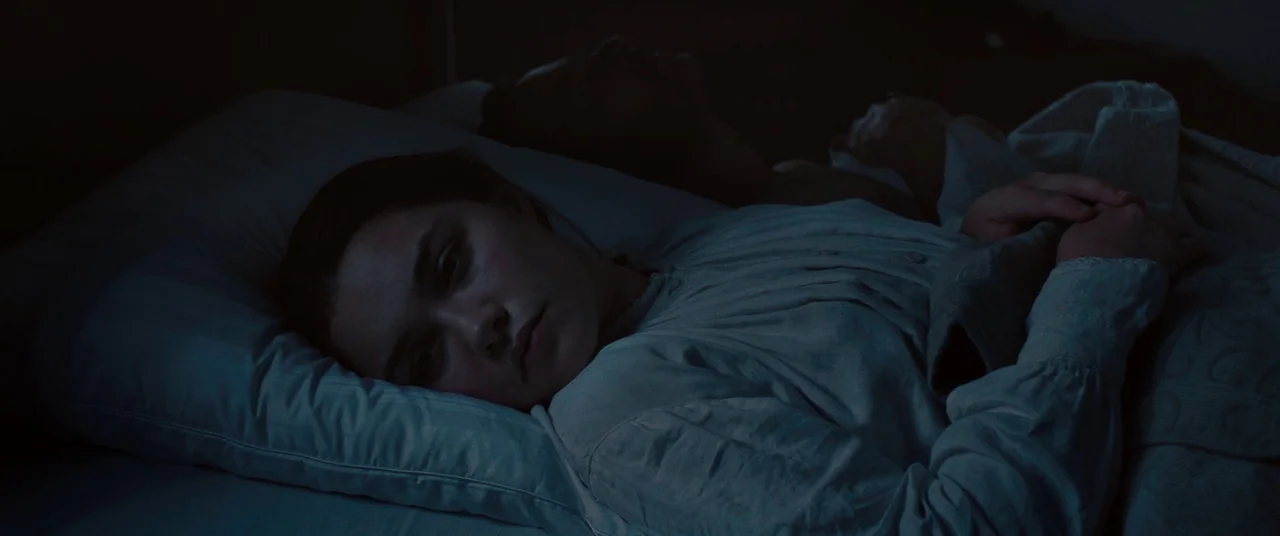 Lady Macbeth (2016) movie still: closeup — A dark, dimly lit bedroom setting features a woman lying in bed. She rests on a pillow with eyes open…