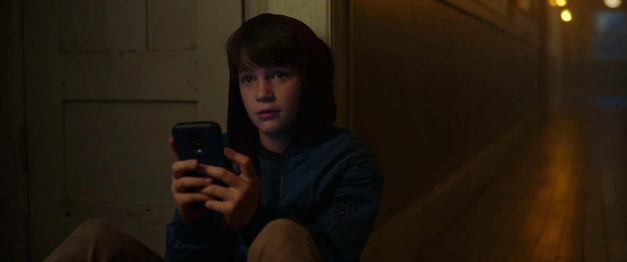 Child's Play (2019) movie still: medium — A dimly lit hallway features a wooden floor and a closed door in the back. A young boy sits on the floor…