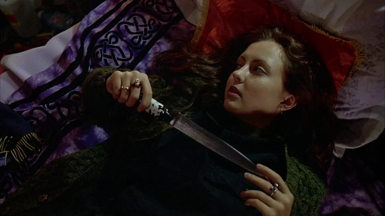 Ginger Snaps (2000) movie still: closeup — A person lies on a bed covered by a purple patterned throw and red pillow. She holds a small, ornate…