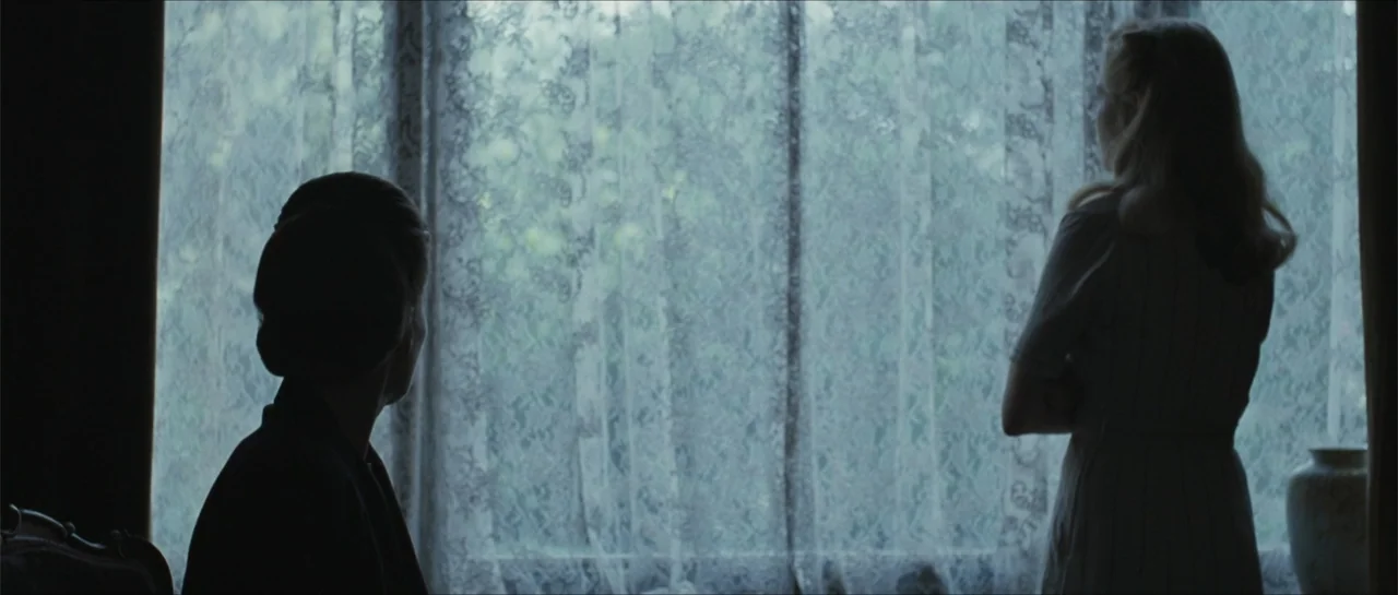 Suite Française (2015) movie still: medium — A dark indoor space features a large window covered by intricate lace curtains. Two women stand in…