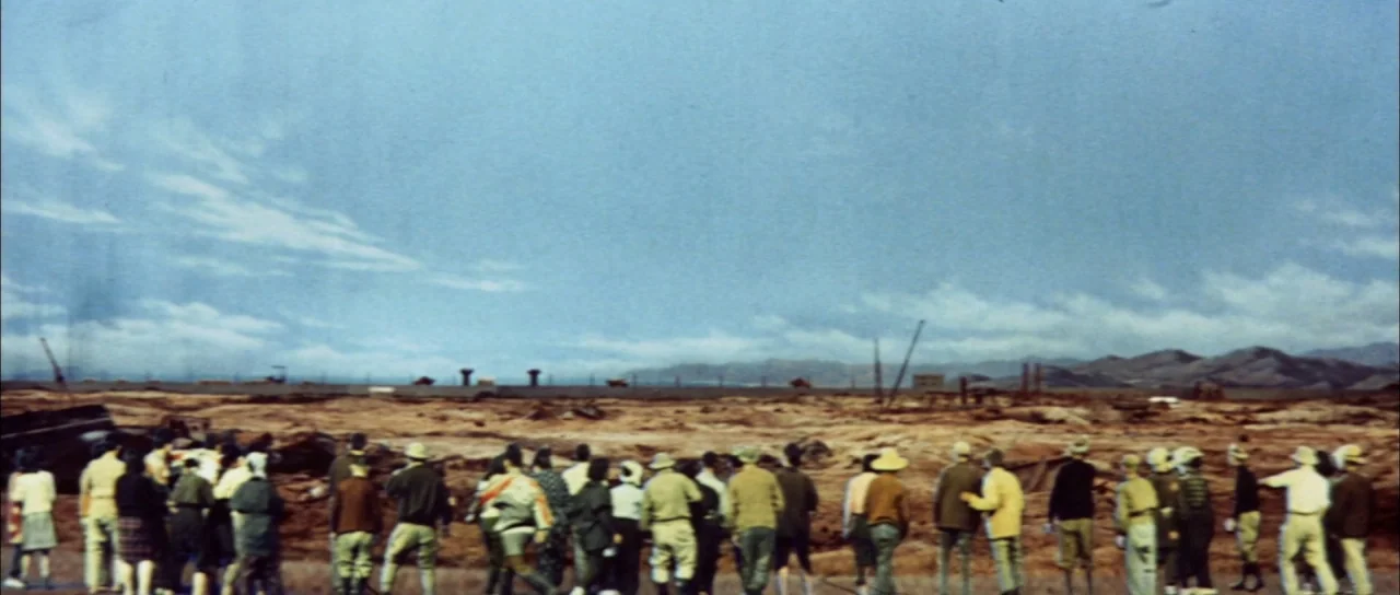 Mothra Vs Godzilla (1964) movie still: wide — A wide, flat landscape features dry, earth-toned terrain under a pale blue sky. A large crowd of…