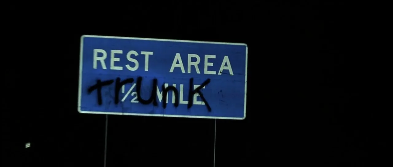Joy Ride (2001) movie still: closeup — A blue highway rest area sign is mounted against a pitch black night sky. Black graffiti spray-painted across…