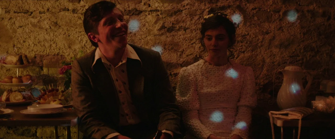 Fréwaka (2024) movie still: medium — A stone-walled interior space features a table spread with plates of food. A laughing man sits beside a woman…