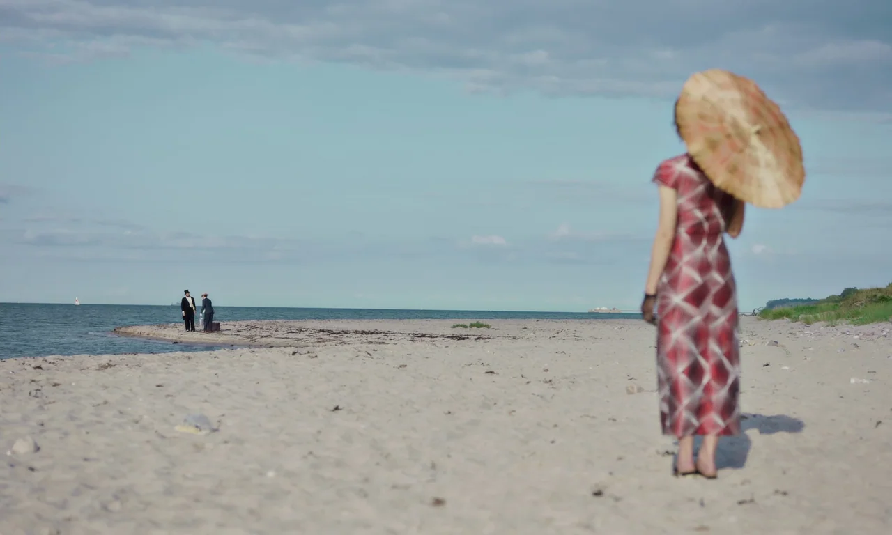Bloodsuckers (2021) movie still: wide — A wide sandy beach stretches toward a calm ocean under a clear blue sky. A woman stands in the foreground…