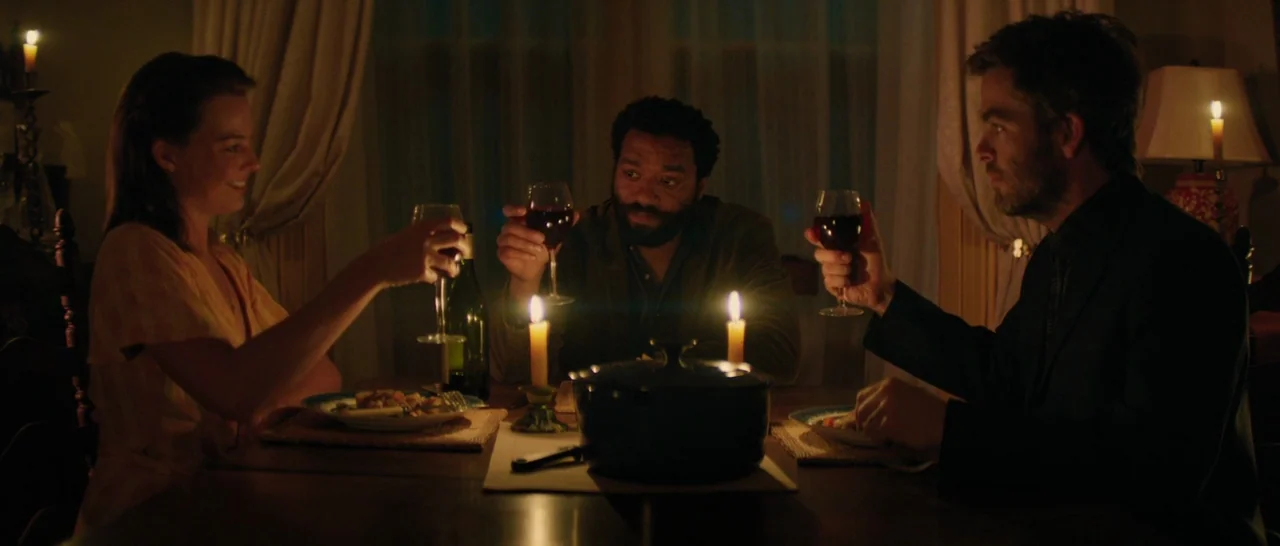 Z for Zachariah (2015) movie still: medium — A dark dining room is lit primarily by candles on the table. Three adults sit together at the wooden…