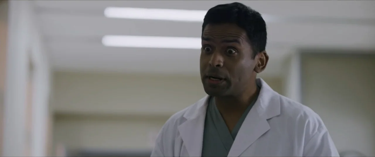 Vicious Fun (2021) movie still: medium — A brightly lit hospital hallway with blurred doors lining the background walls. A man in medical scrubs and…