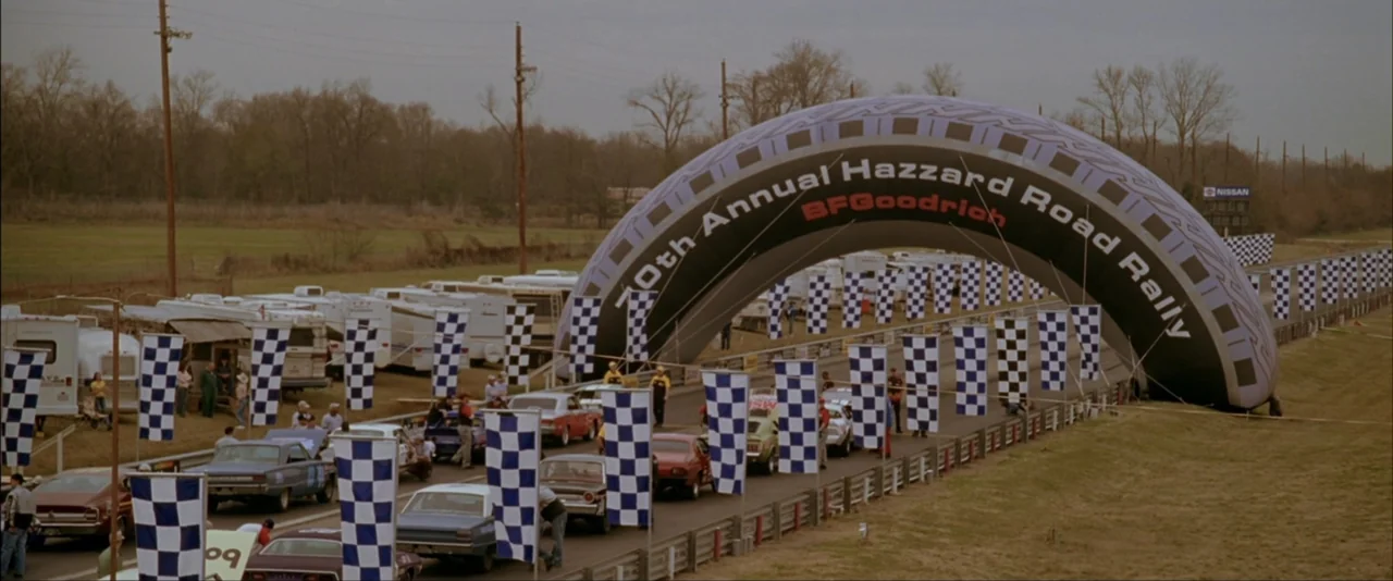 The Dukes of Hazzard (2005) movie still: wide — An outdoor racing rally features a large inflatable arch over a paved road. Multiple vintage…