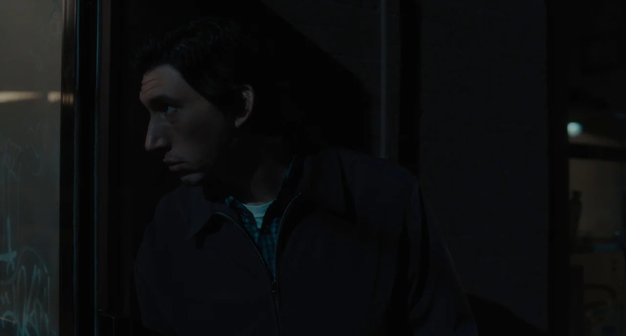 Paterson (2016) movie still: medium — A dark outdoor setting at night features a man standing near a doorway. The man looks through a glass pane…