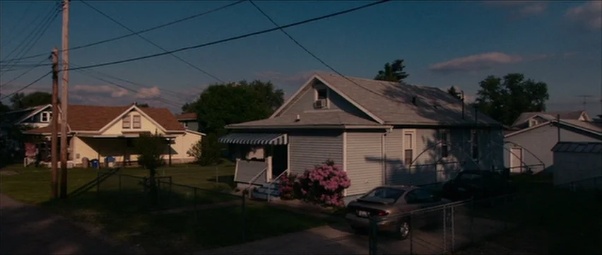 Bubble (2005) movie still: establishing — A quiet suburban street features several small houses with light-colored siding and yards. Power lines…