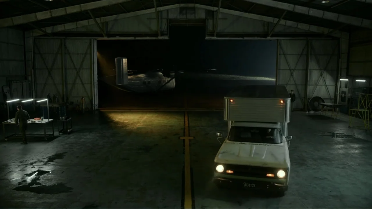 Transfers (2024) movie still: wide — A dark, spacious airplane hangar contains a workbench and a large open bay door. A box truck drives toward the…