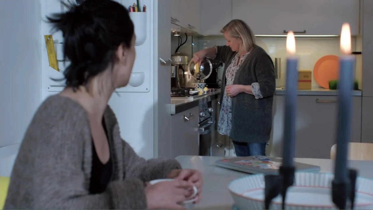 Code of Trust (2019) movie still: medium — A residential kitchen with modern cabinetry and a dining table in the foreground. Two women interact in a…