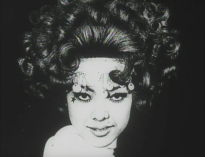 Funeral Parade of Roses (1969) movie still: closeup — A dark environment features a performer with elaborate hair and headpiece against black. The…