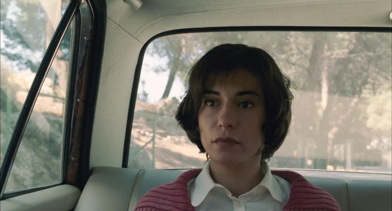 Incendies movie still: medium — The interior of a vintage car with light-colored upholstery and dark window frames. A woman sits in the passenger…