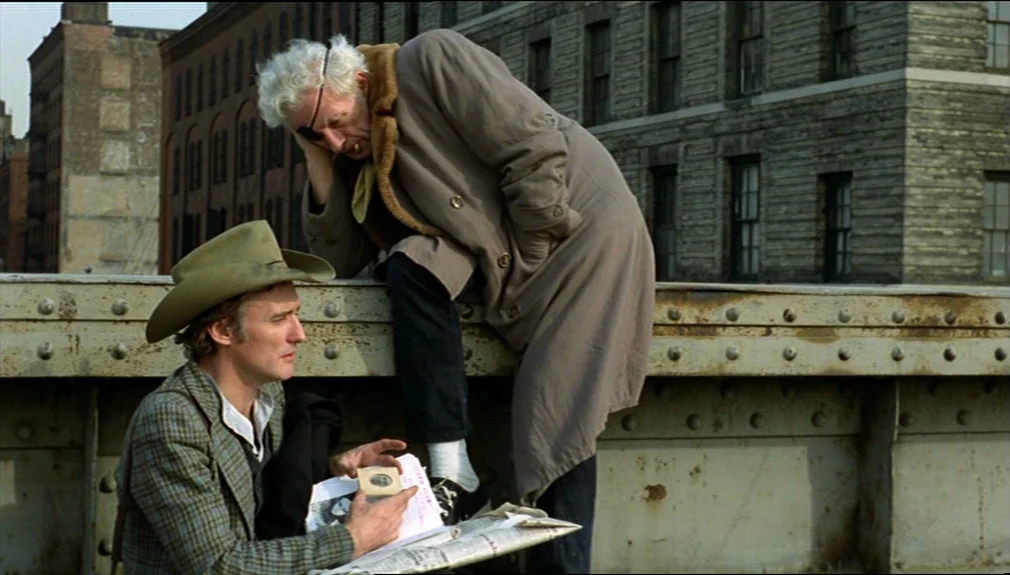 The American Friend (1977) movie still: medium — A metal bridge structure overlooks industrial brick buildings under a clear daytime sky. A younger…
