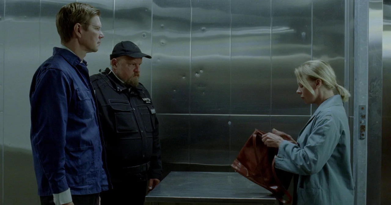 Fallen Leaves (2023) movie still: medium — A sterile security room features metallic walls with dents and a stainless steel counter. A woman stands…