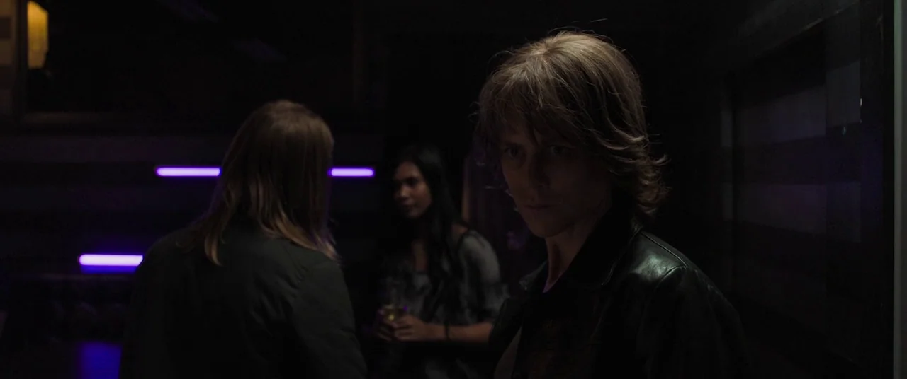 Destroyer (2018) movie still: medium — A dimly lit lounge space with purple neon lines illuminating a dark textured wall. A man in a leather jacket…