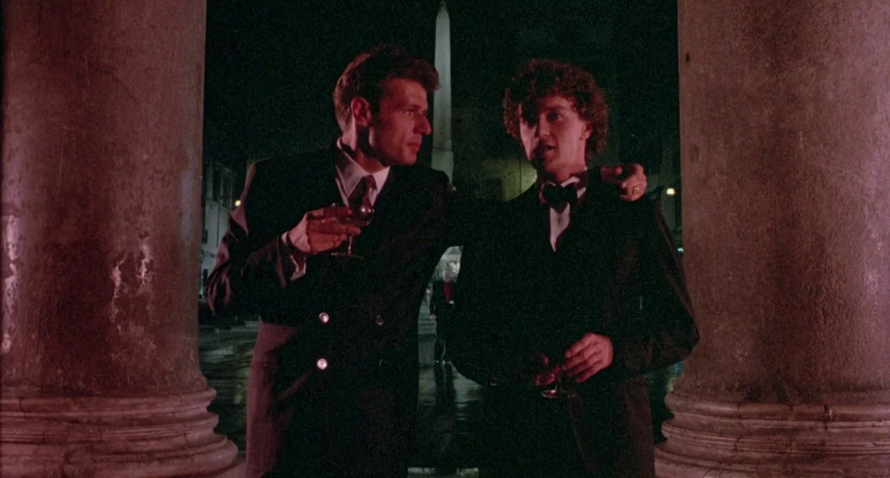 The Belly of an Architect (1987) movie still: medium — Two men in formal attire stand together between two large stone columns at night. An obelisk…