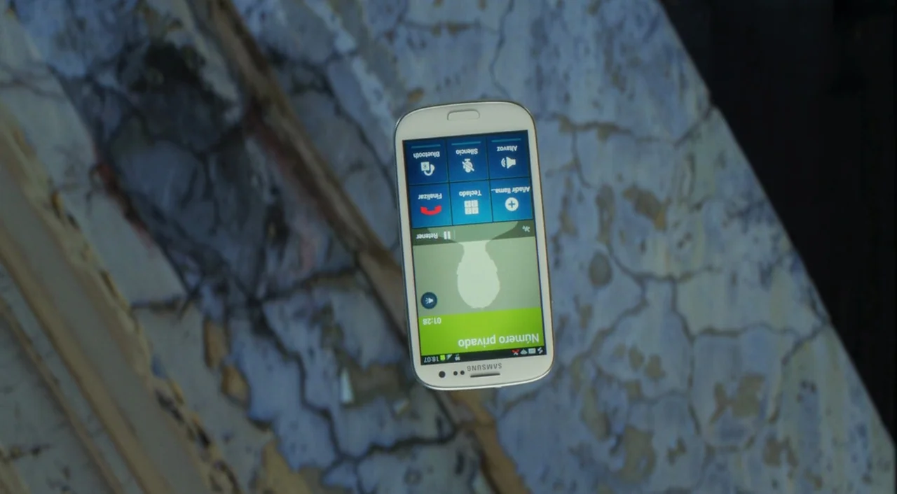 I’m So Excited! (2013) movie still: closeup — A white Samsung smartphone floats against a textured background of weathered and cracked concrete. The…