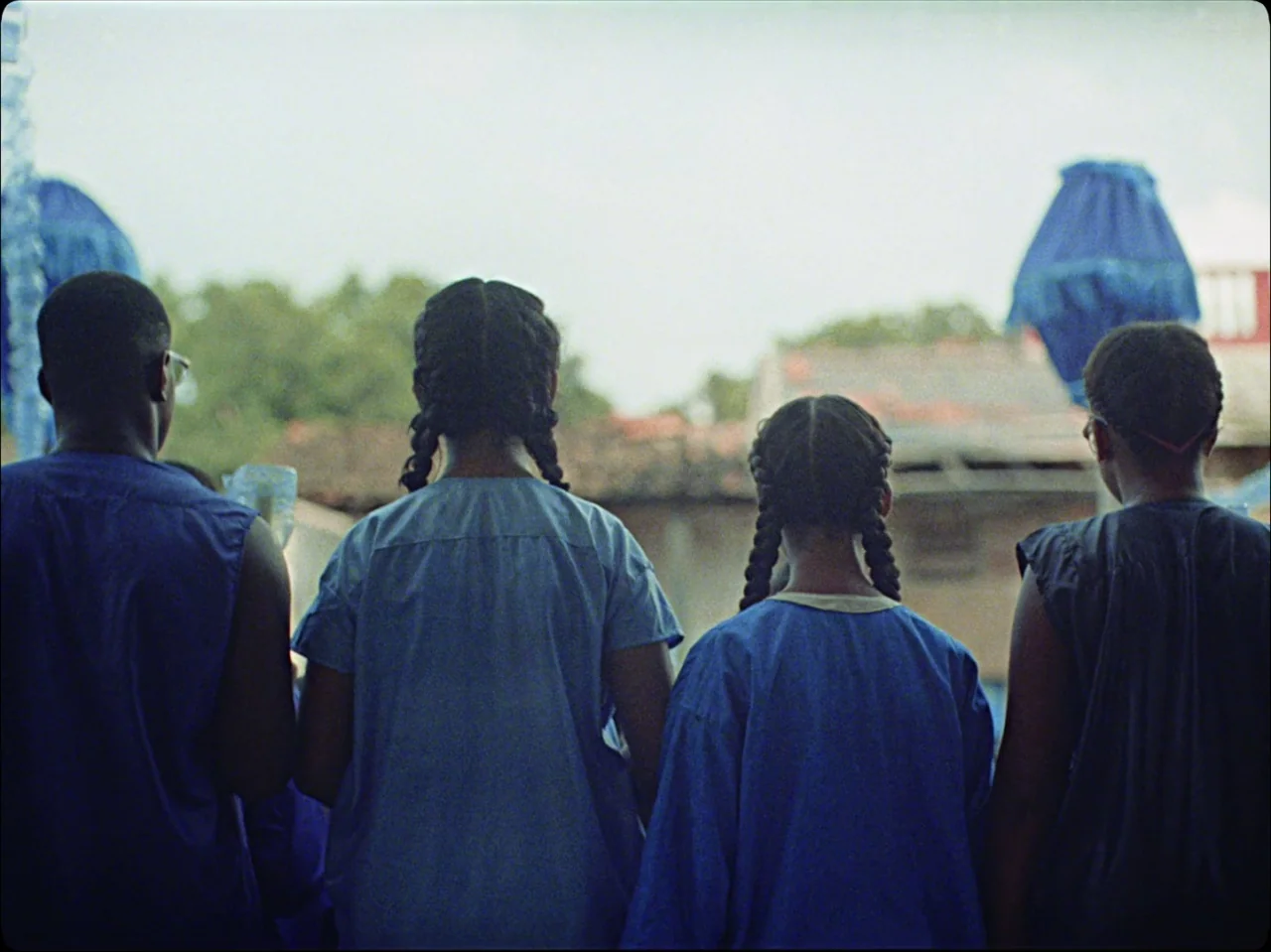 Guava Island (2019) movie still: medium — Four people stand together in a row against an outdoor background with rustic structures. The individuals…