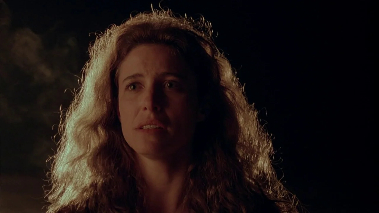 The Rapture (1991) movie still: closeup — A woman stands in darkness, illuminated by a warm, flickering light source. She looks toward the side of…