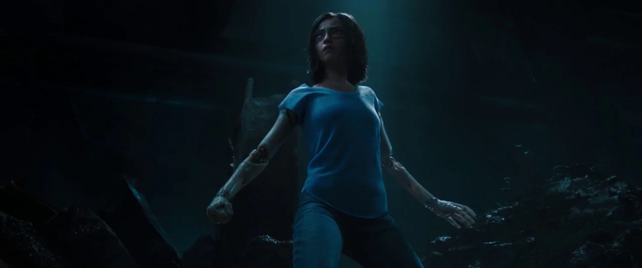 Alita: Battle Angel movie still: medium — A dark, desolate junkyard is filled with piles of rusted metal and debris. A young girl with robotic arms…