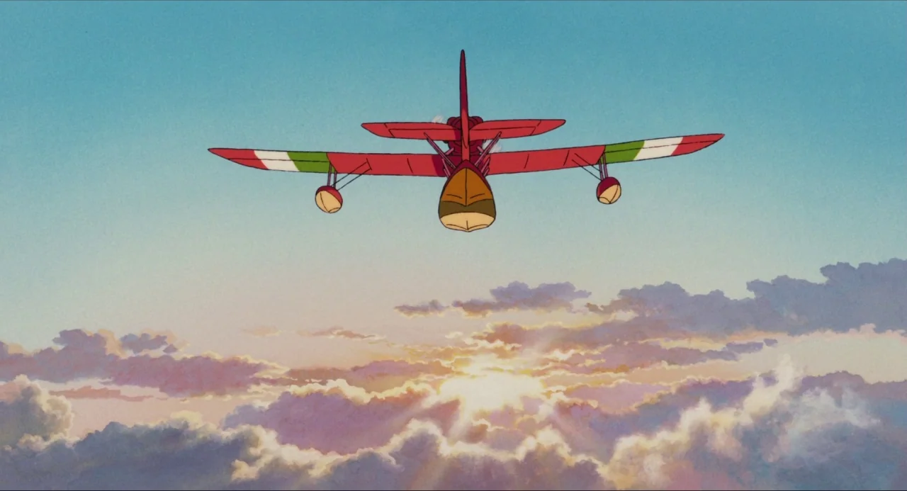 Porco Rosso (1992) movie still: wide — A bright red seaplane flies above a layer of sunset clouds. The sky fades from light blue to a pale horizon…