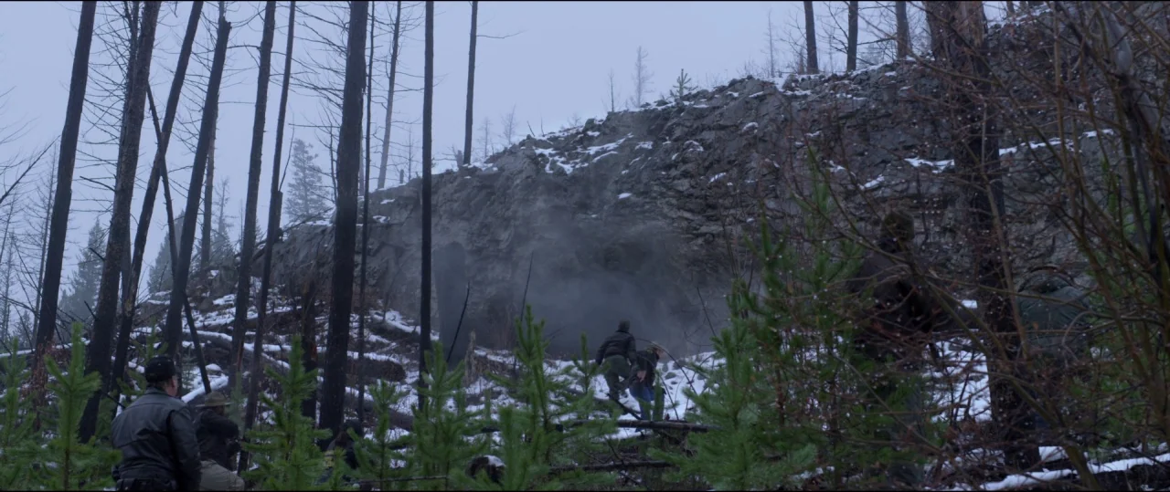 Buster’s Mal Heart (2016) movie still: wide — A snowy, sparse mountainside features burnt trees and a large, dark rock formation. Several…