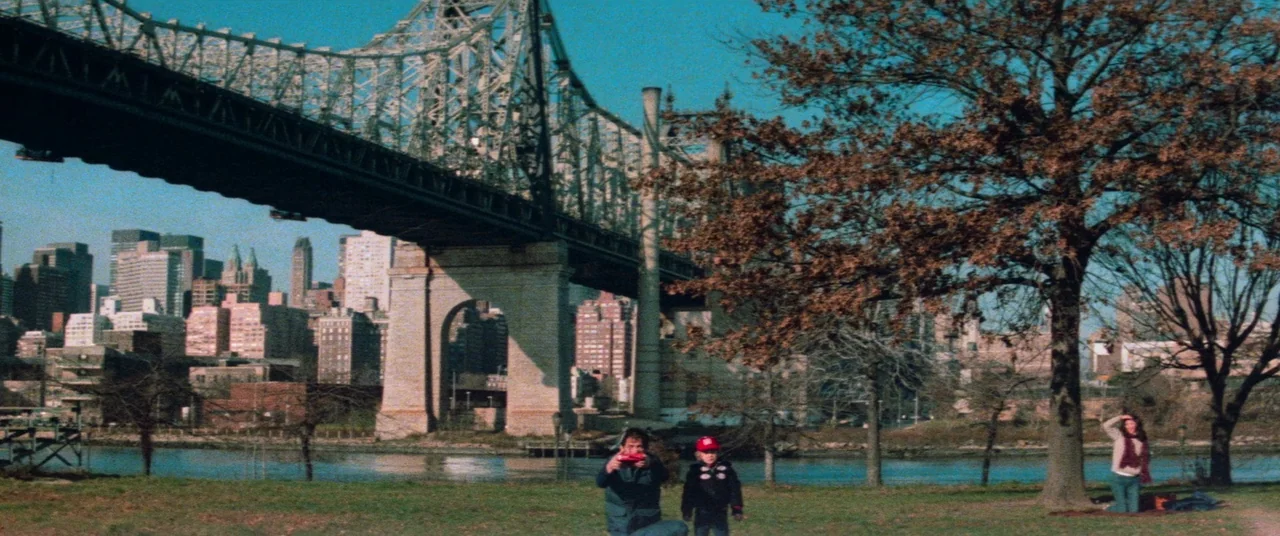 Vigilante (1982) movie still: wide — A riverside park features a massive steel truss bridge spanning across the water. Three people occupy the…