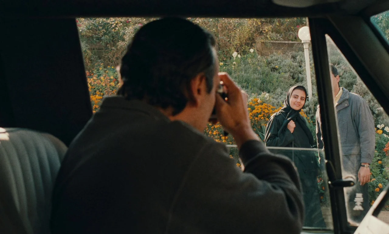 Taste of Cherry (1997) movie still: medium — A man sits inside a vehicle while aiming a camera through the open door. Two people stand outside in a…