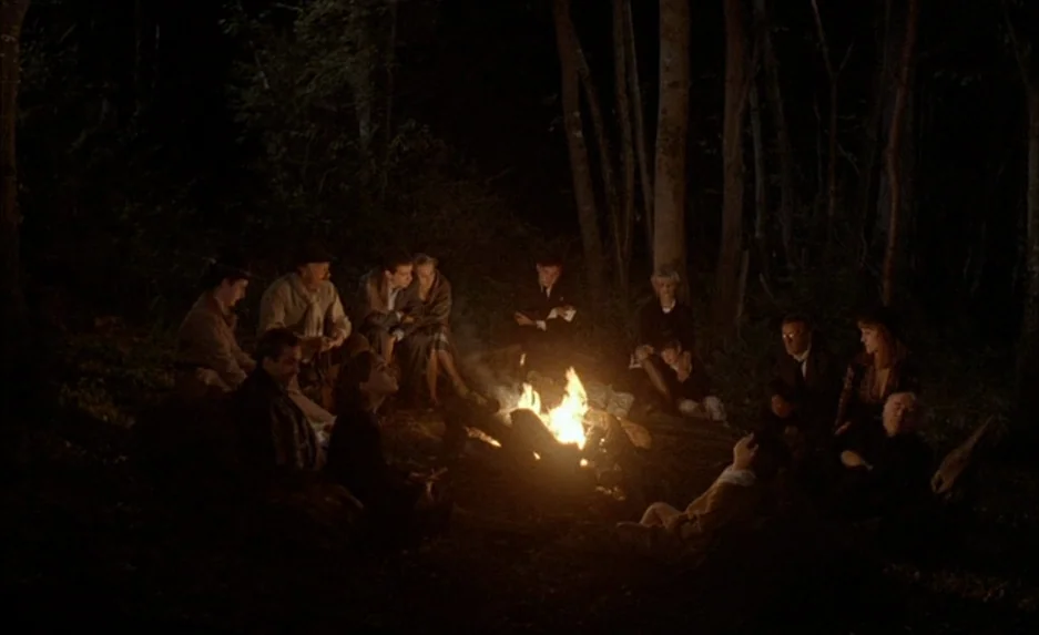 Milou en Mai (1990) movie still: wide — A group of people sits on the ground in a dark, wooded clearing at night. A bright, central campfire…