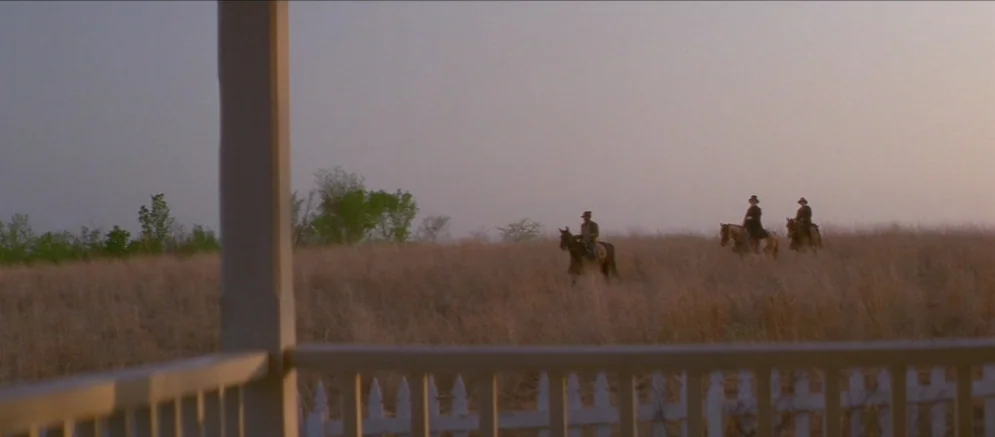 Ride With The Devil movie still: wide — An open expanse of dry golden grass stretches towards a treeline under a hazy sky. Three men on horseback…