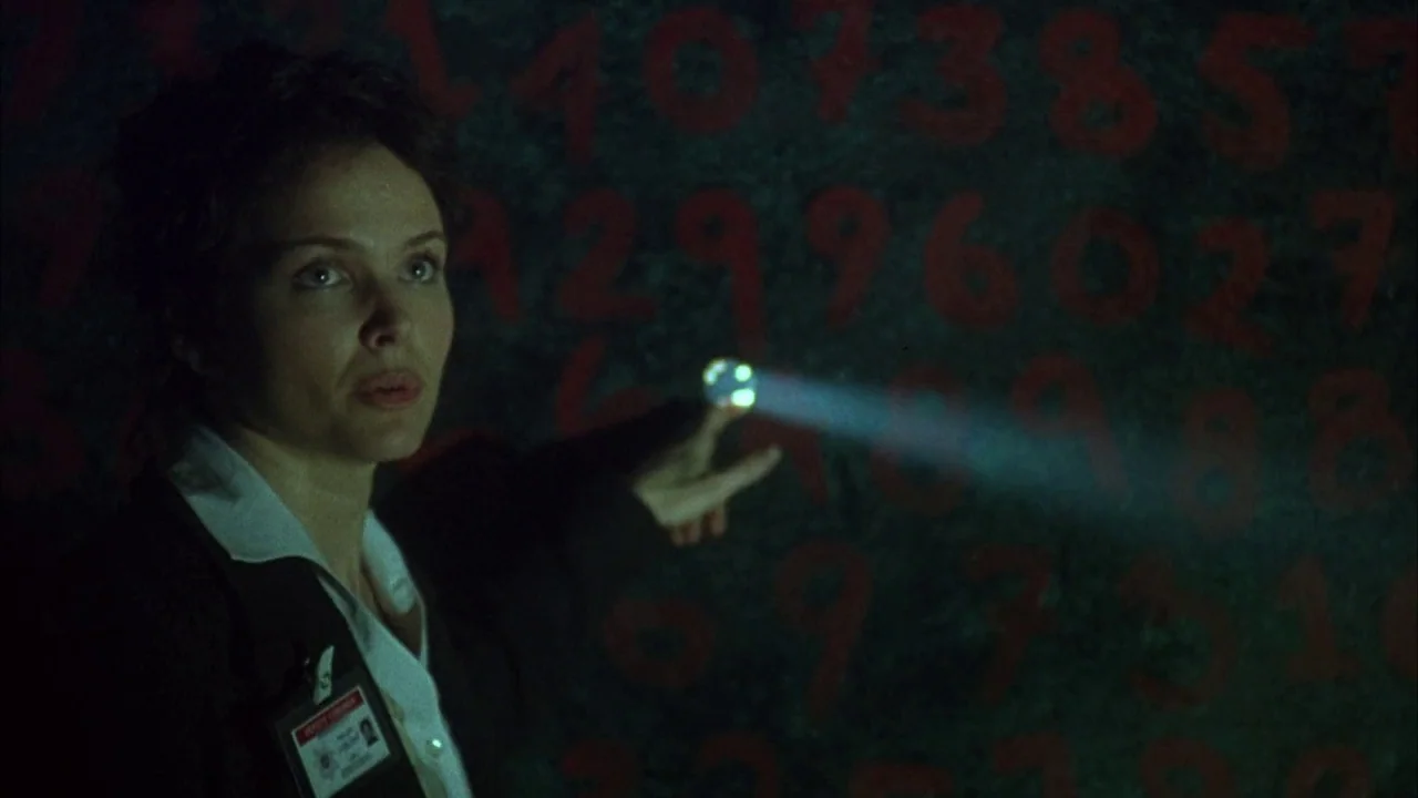 Saw movie still: medium — A dimly lit room with walls covered in large red painted numerical sequences. A woman inspects the numbers using a small…