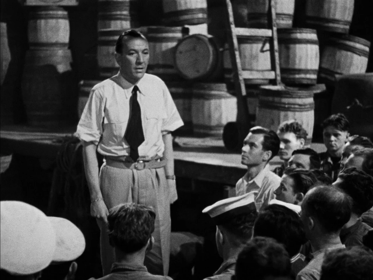 In Which We Serve (1942) movie still: medium — A dimly lit storage warehouse features wooden barrels stacked high in the background. A man stands…