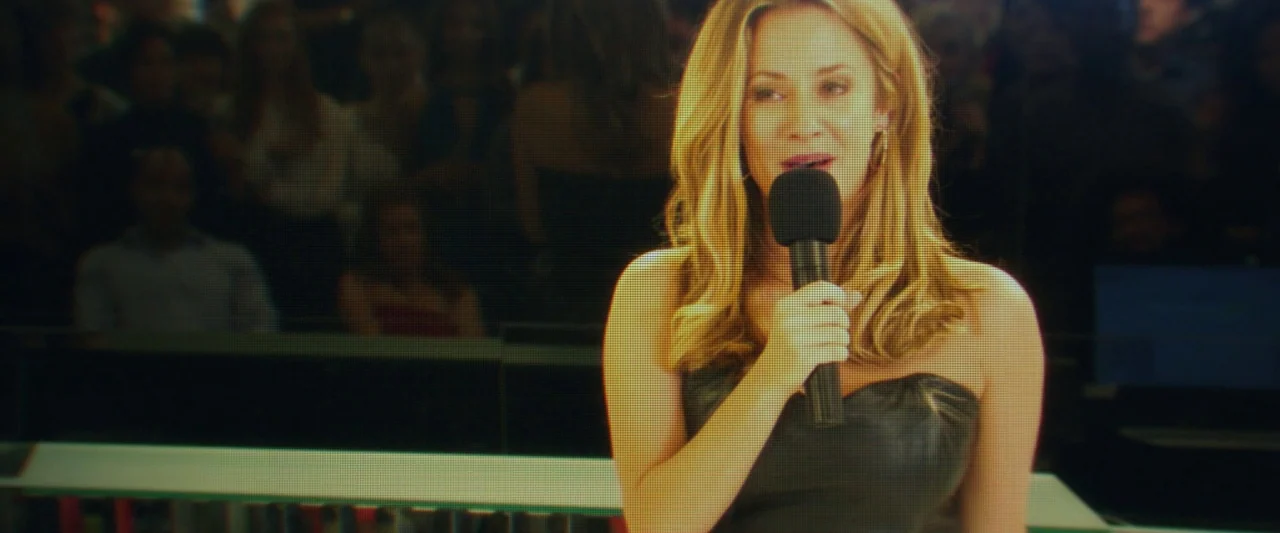Greed (2019) movie still: medium — A television studio environment with a blurred audience seated in the background. A woman holds a black…
