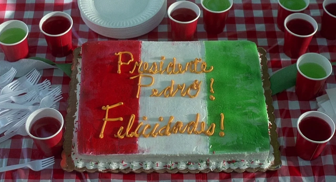 Napoleon Dynamite (2004) movie still: closeup — A rectangular party cake decorated with red, white, and green icing sits on a table. Red plastic…