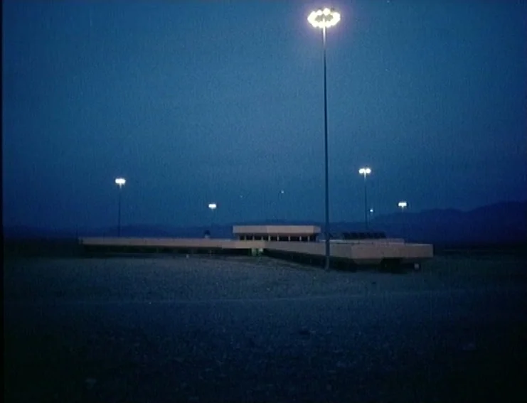 Ghosts… Of The Civil Dead (1988) movie still: wide — A flat-roofed building sits in a dark, expansive area under a night sky. Tall, illuminated…