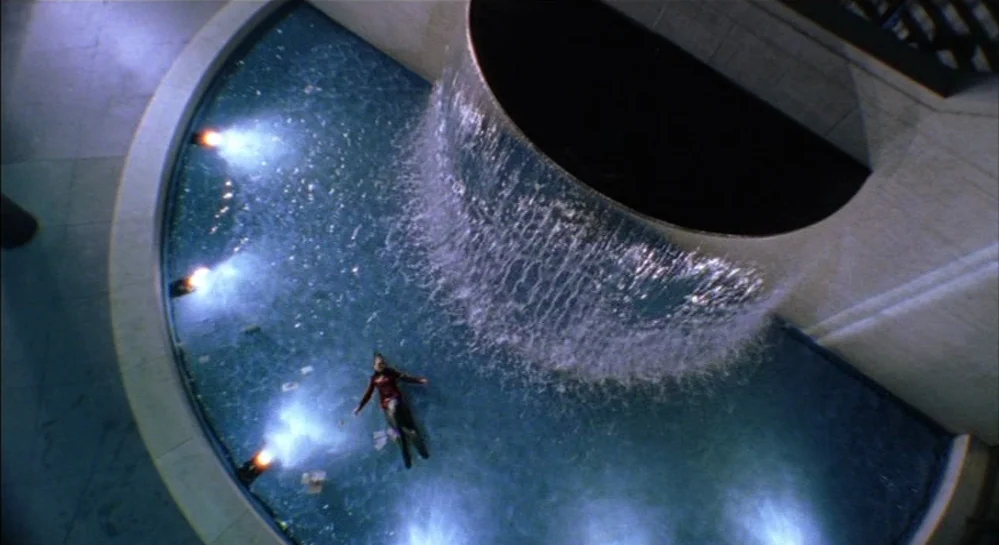 Hamlet (2000) movie still: wide — A semi-circular indoor fountain features a waterfall cascading over a smooth curved stone wall. A person wearing a…