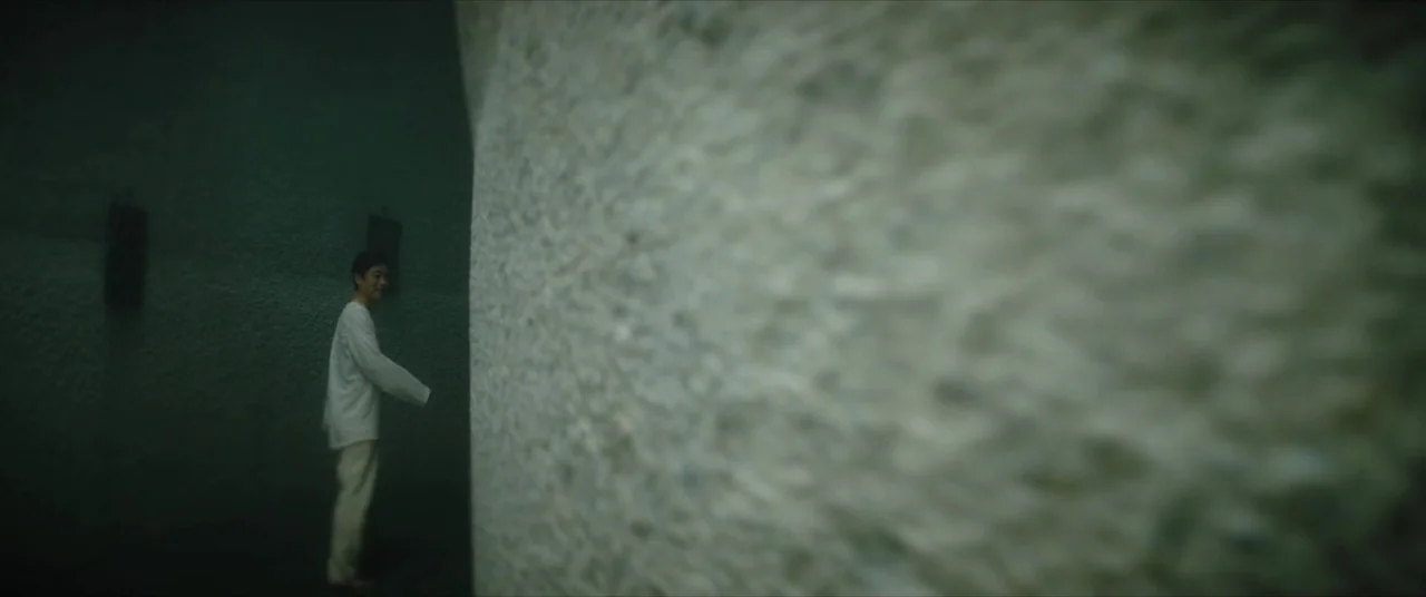 The Divine Fury (2019) movie still: wide — A narrow corridor features a rough, textured white stone wall on the right side. A man wearing…