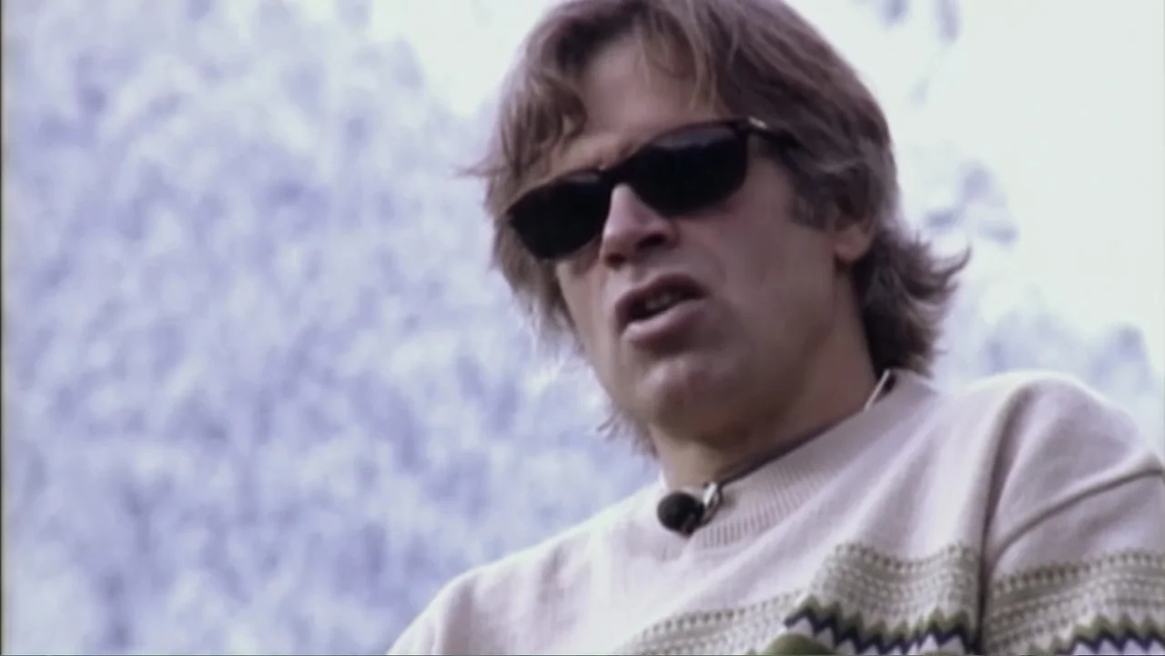 Dear Rider: The Jake Burton Story (2021) movie still: closeup — A man stands outdoors in front of a blurry, light-colored mountainous landscape. The…